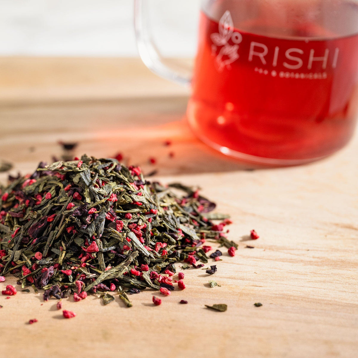 A pile of Raspberry Green Tea leaves next to a cup of Rishi Tea & Botanicals.