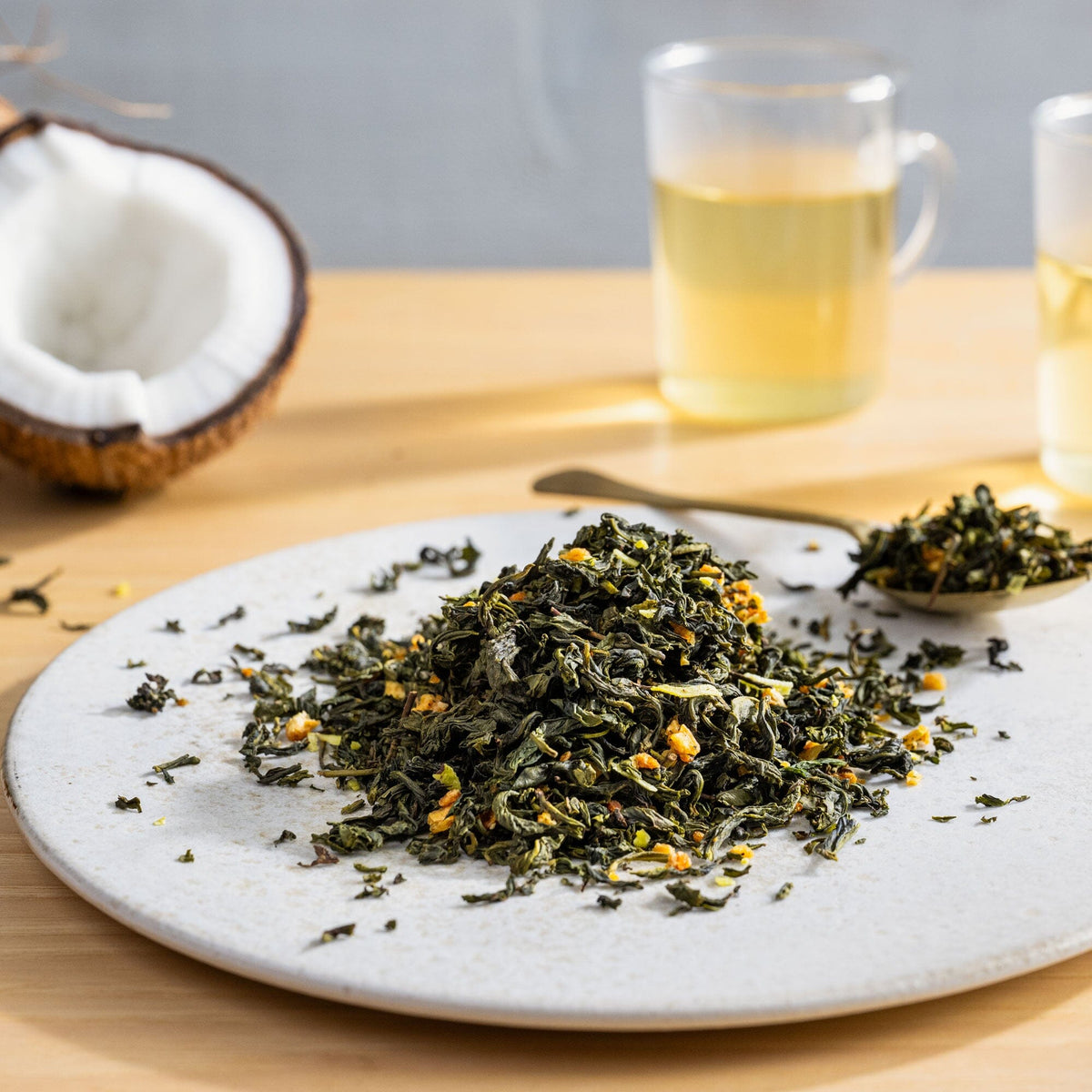 The table features a plate with Rishi Tea & Botanicals' Tropical Coconut Oolong loose leaf tea, a spoonful of the oolong tea, a halved creamy coconut, and two glass mugs of the tropical blend.