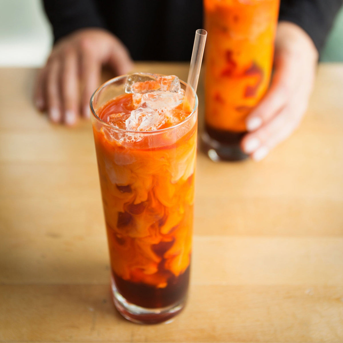 A glass of iced Thai tea, crafted with Sweet Thai Tea Powder from Rishi Tea & Botanicals and milk, swirled on a wooden table. In the background, another glass is being placed by someone.