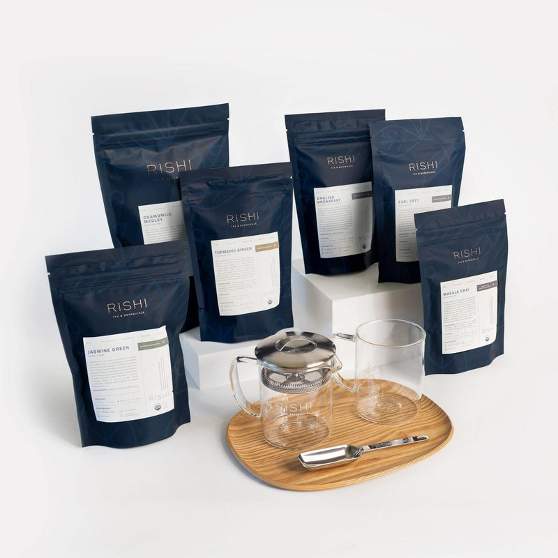 A Rishi Tea & Botanicals bundle with six tea blends, a glass teapot with metal infuser, two glass cups, and a metal scoop on a wooden tray—all essential brewing tools shown against a white background.