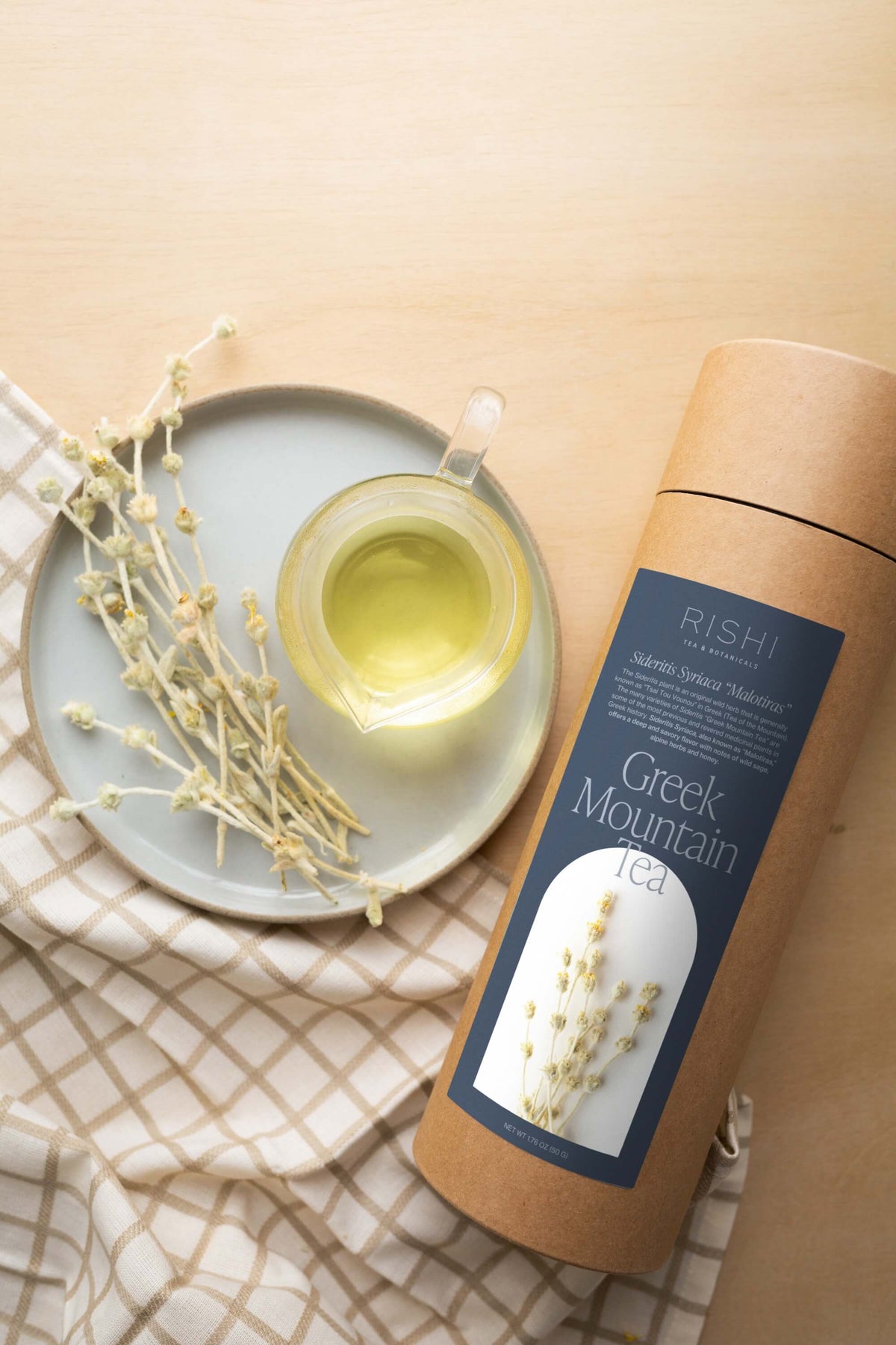 A cylindrical container of Rishi Tea & Botanicals Greek Mountain Tea Malotiras with two small bowls of the botanical tea and dried herb stems on a plate, placed on a checkered cloth, next to a glass pitcher of the nectar-like flavor.