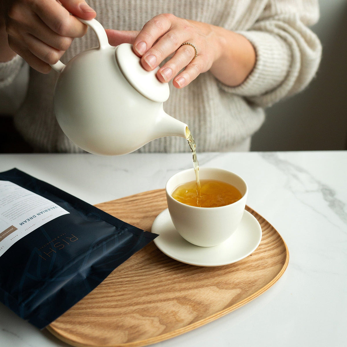 A person gracefully pours tea from a Kinto Pebble Porcelain Teapot, crafted by Kinto USA, Inc, featuring a drip-free spout, into a cup on a wooden tray, with the aroma mingling from the nearby tea package.