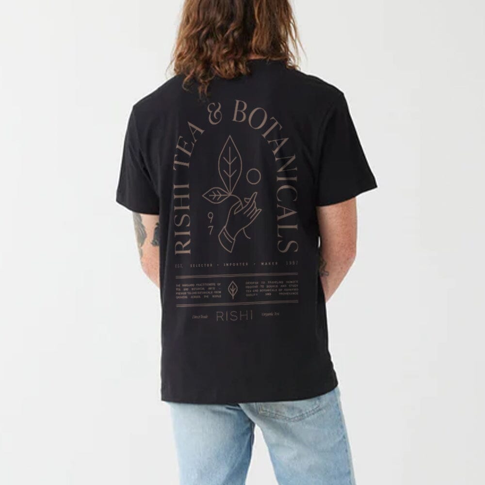 Sporting the Rishi Weekend T-Shirt - Black, adorned with the "Rishi Tea & Botanicals" logo and a design on the back illustrating a hand holding a plant. Crafted from organic cotton and available in unisex sizing, this shirt upholds Fair Trade principles.