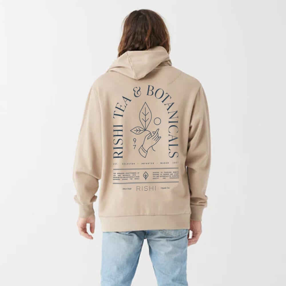 A person in the Rishi Everyday Hoodie - Tan, featuring a "Rishi Tea & Botanicals" design on the back and made from ultra-soft organic cotton fleece, stands facing away from the camera against a plain background.