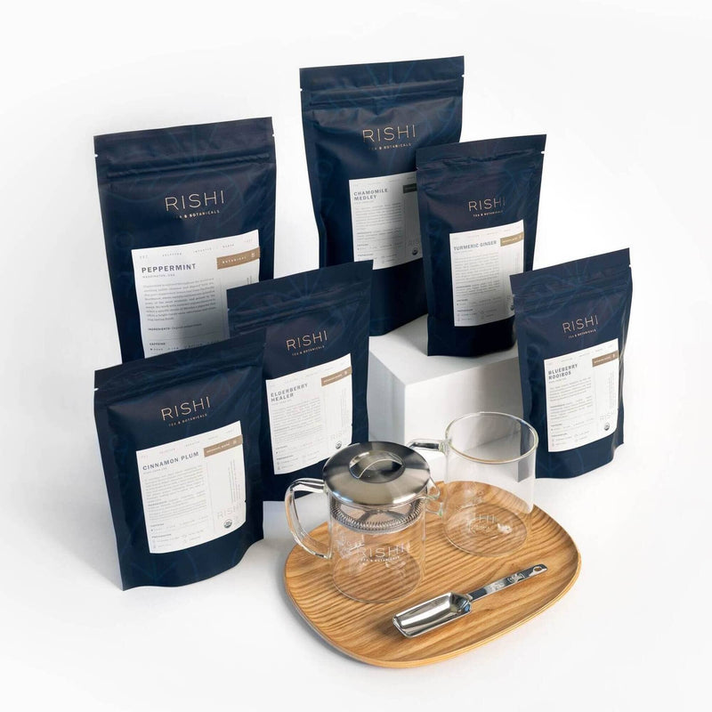 Rishi Tea & Botanicals herbal tea bags, a glass teapot, two glass cups, premium teaware, a metal scoop, and a wooden tray are arranged on a white surface.