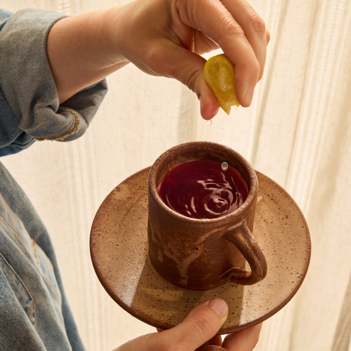 A person squeezes a lemon wedge over a brown ceramic cup filled with a dark red Rishi Tea & Botanicals infusion, holding the cup on a matching saucer.