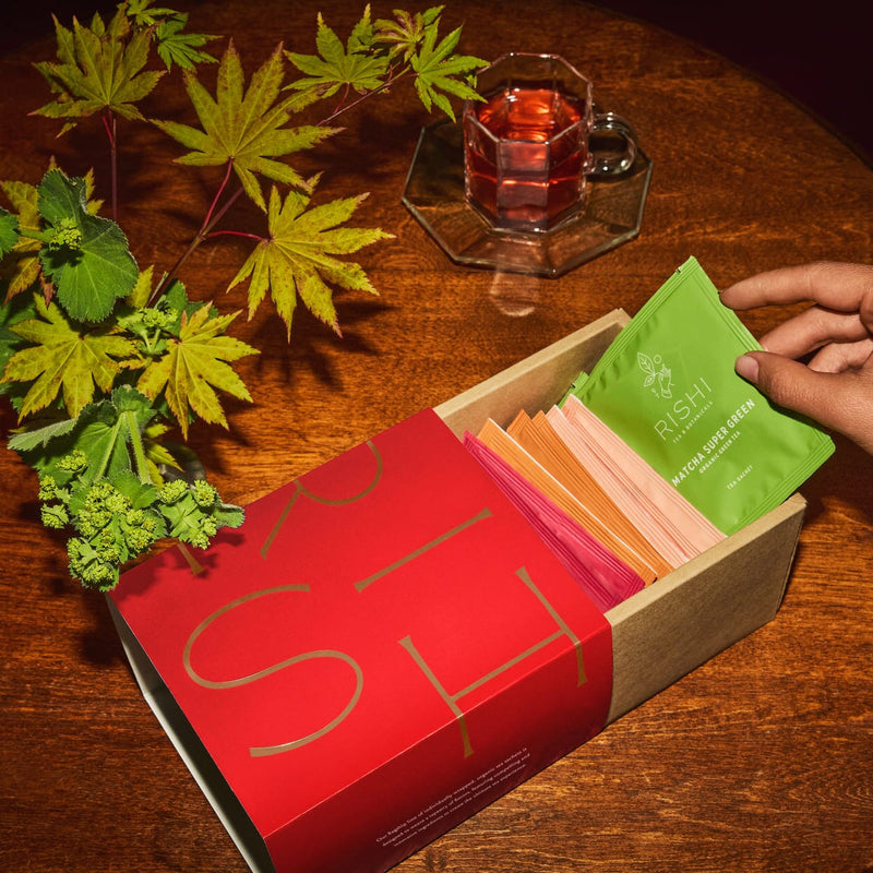 A hand selects a green Rishi tea packet from a box of assorted tea sachets on a wooden table with a leafy branch and a glass cup of tea.