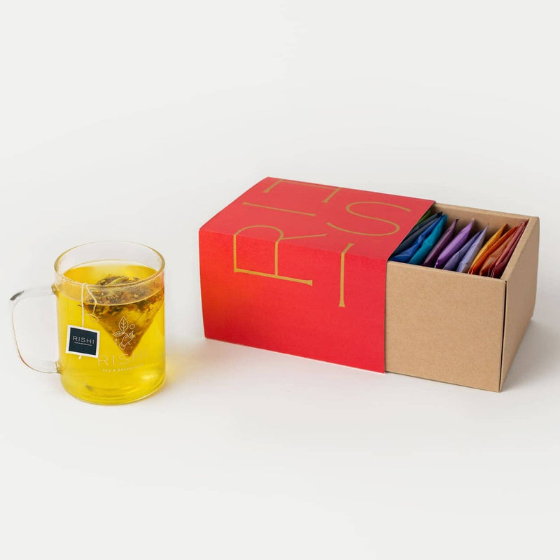 A glass mug of tea sits next to a red and brown box containing assorted colorful tea sachets arranged in rows.