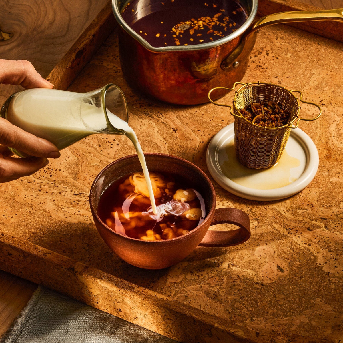 A hand pours milk from a glass jar into a brown cup of Rishi Tea & Botanicals chai on a wooden tray, with a pot and a tea strainer nearby. Hints of spicy ginger linger in the air, promising a warming and aromatic experience.