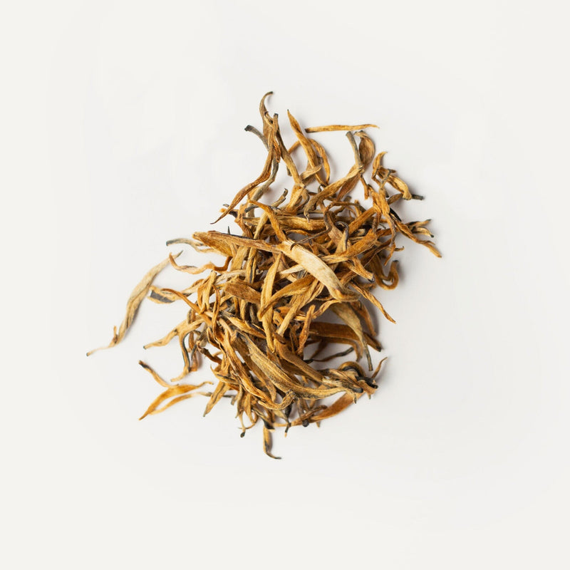 A pile of loose, dried Yunnan Dian Hong Golden Buds tea leaves with a mix of golden and brown hues, sourced from an organic tea garden, displayed on a plain white background by Rishi Tea & Botanicals.