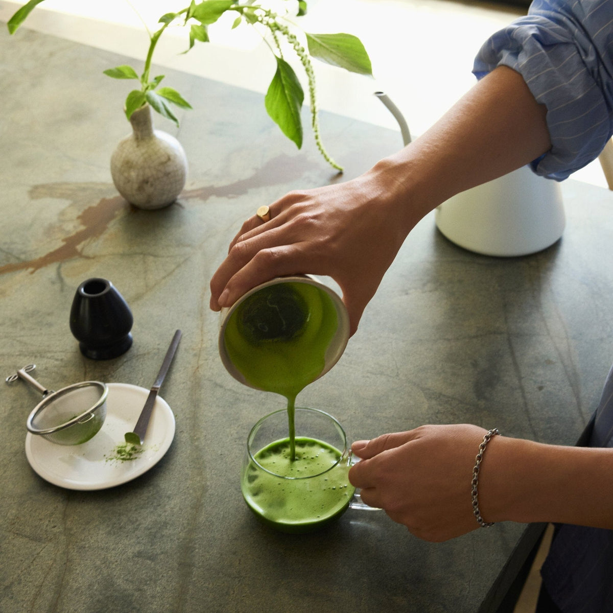 A person pours vibrant green Rishi Tea & Botanicals organic matcha tea from a small pitcher into a glass cup on a kitchen counter, with a strainer and a small plate nearby.