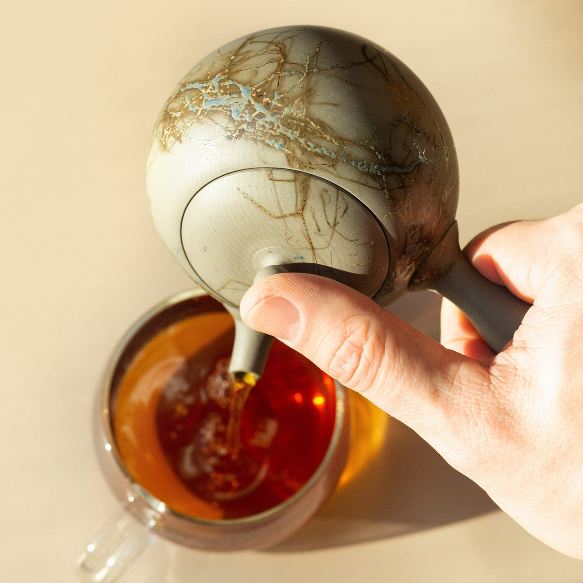 A hand pours tea from a round Tokoname kyusu by Chato Co., Ltd. into a clear glass cup of amber tea, highlighting the teapot's elegant clay strainer.