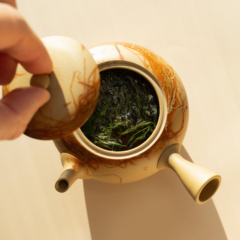 A hand lifts the lid of a Chato Co., Ltd. Tokoname kyusu, revealing steeped green tea leaves inside this limited-edition teapot.