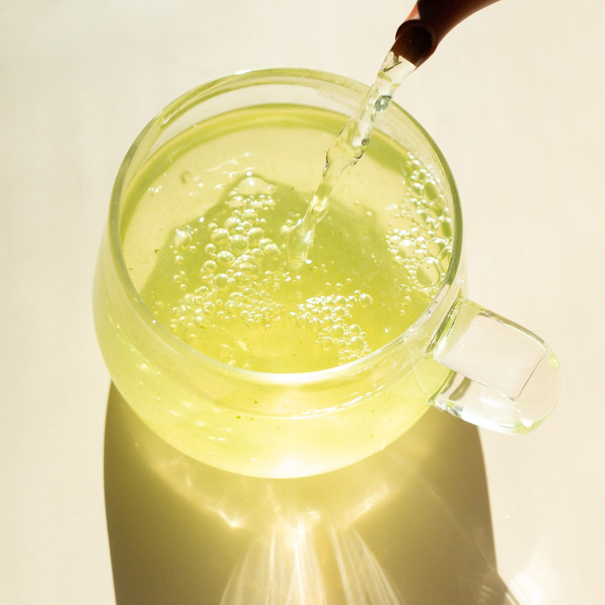 A clear glass cup from Rishi Tea & Botanicals is filled with light yellow, bubbly Japanese tea being poured from above, creating a shadow on a light surface.