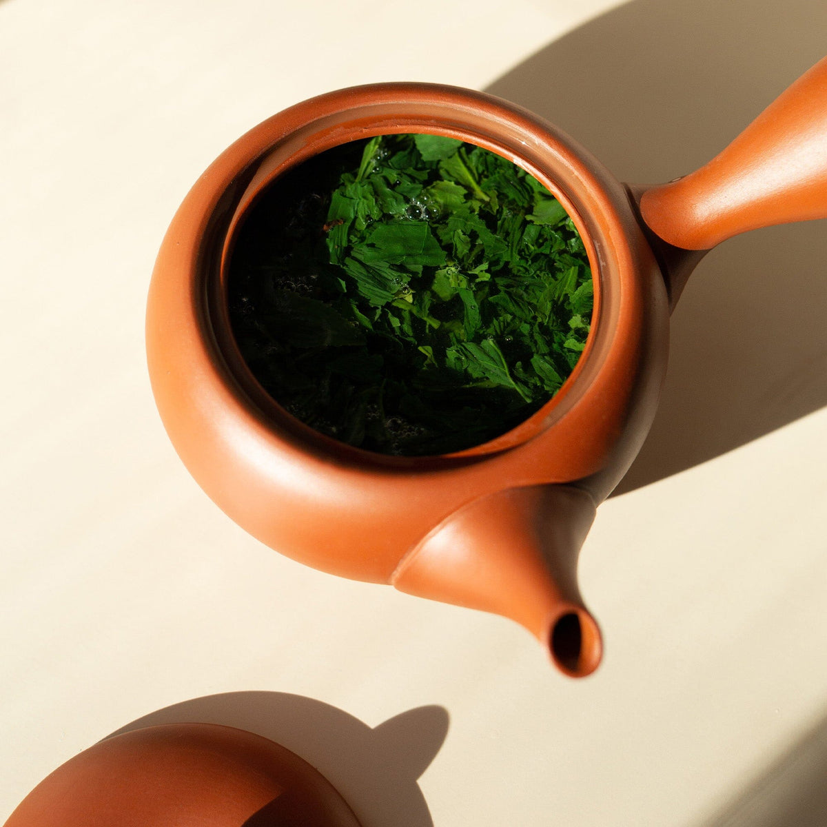 A brown teapot from Rishi Tea & Botanicals, filled with vibrant gyokuro green tea leaves, sits open on a light surface, photographed from above to highlight the fresh color and rich umami aroma of this competition-grade tea.