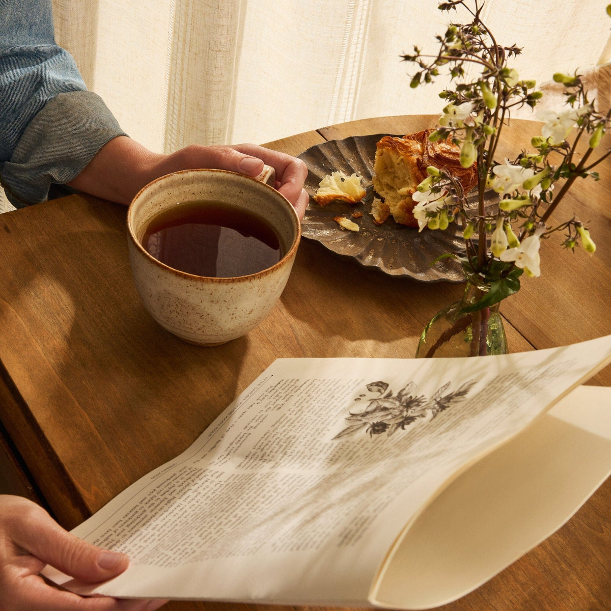 A person holds an open book at a wooden table, accompanied by a cup of creamy black tea from Rishi Tea & Botanicals, a plate with pastry, and a small vase of flowers.