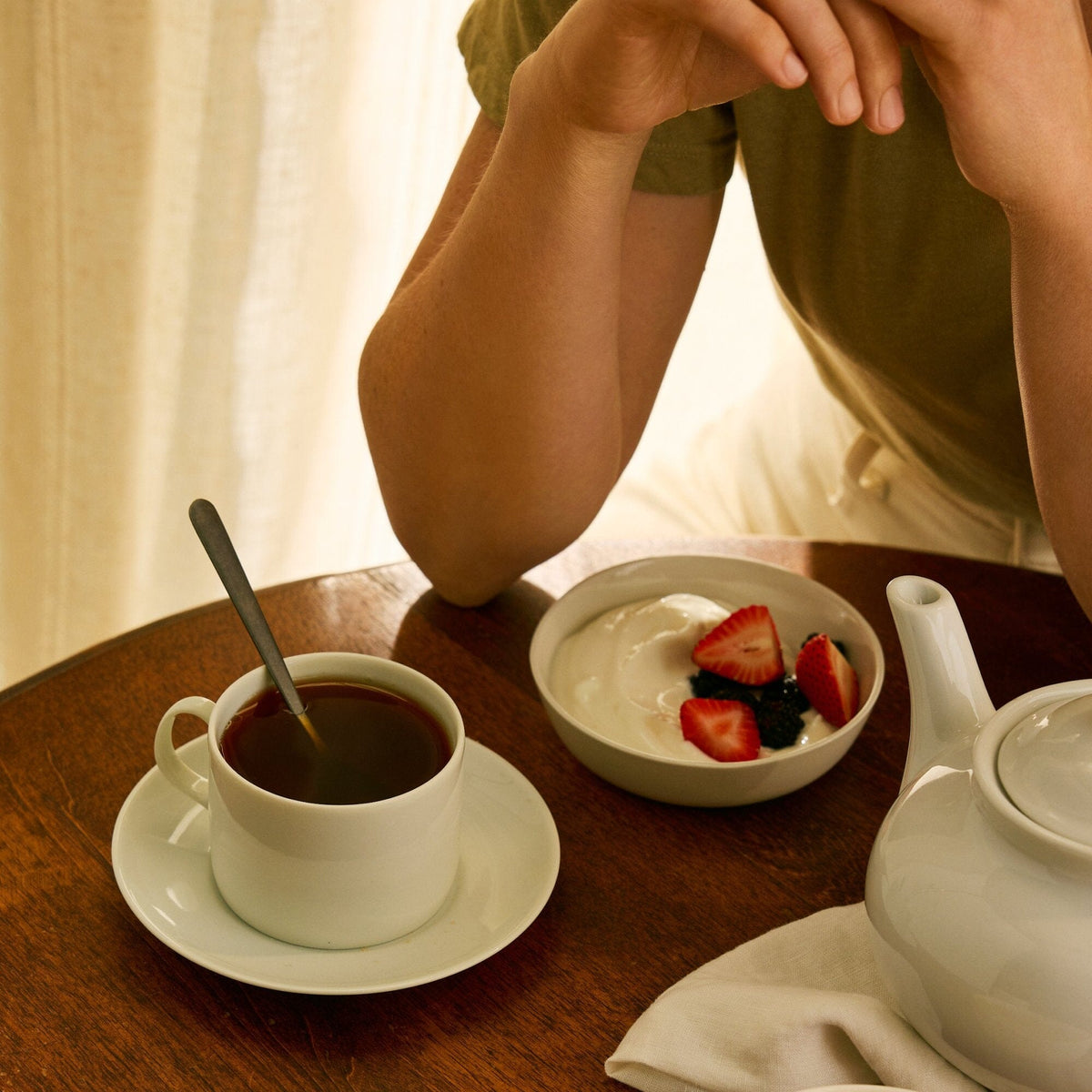 A person sitting at a table with a cup of Rishi Tea & Botanicals adventurous black tea, a teapot, and a bowl of yogurt topped with strawberries and blueberries.