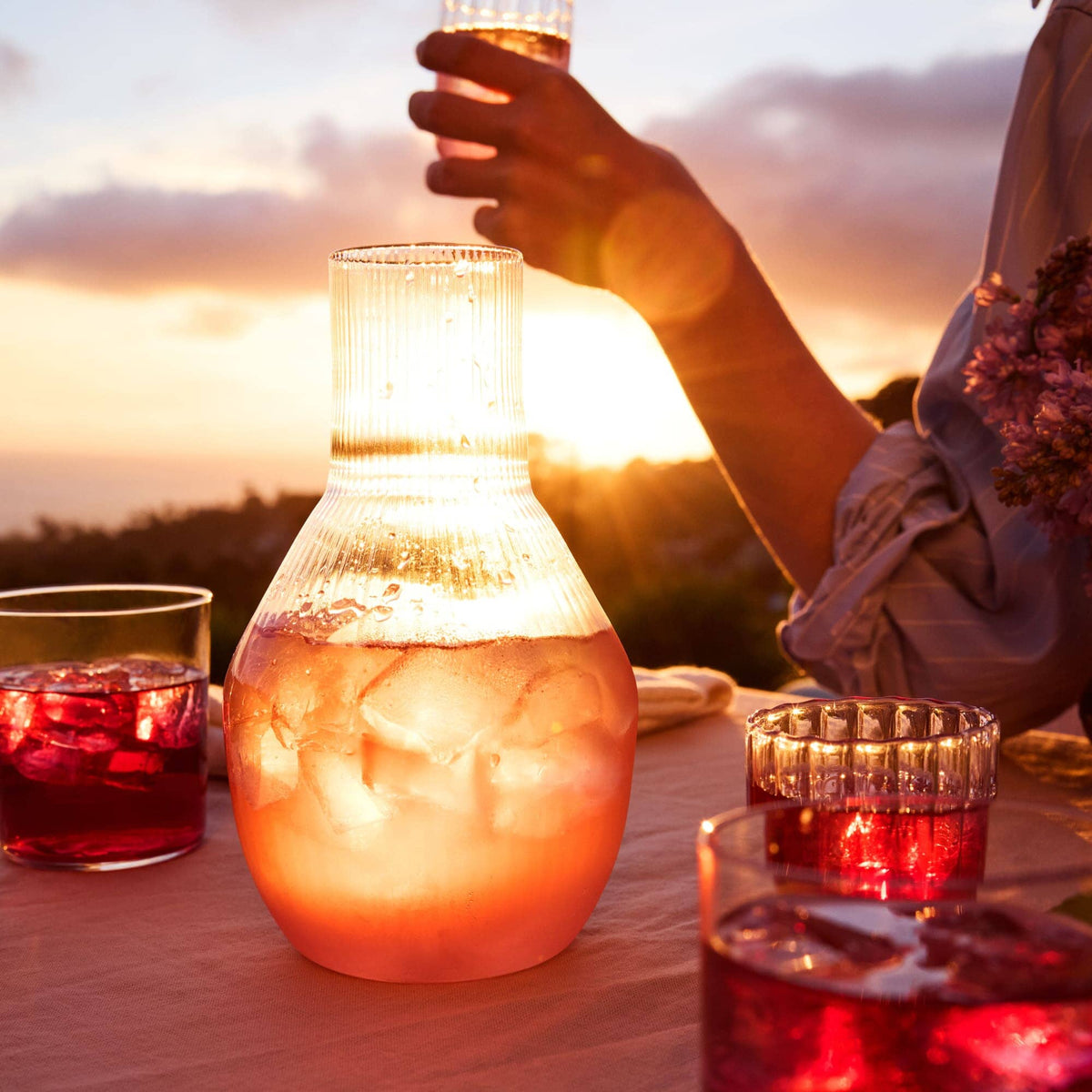A glass pitcher with a reddish yuzu-flavored drink from Rishi Tea & Botanicals sits on a table with matching glasses at sunset, while someone holds an iced tea in the background.